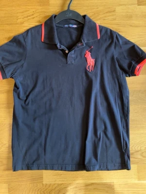 Svart pikétröja med röd logga - Klassisk svart pikétröja från Polo Sport med stor röd broderad logga på bröstet och röda detaljer på krage och ärmslut. Tröjan har korta ärmar och knappar vid halsen. Perfekt till jeans eller shorts för en sportig look.