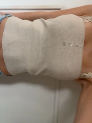 Beige ribbad tubtopp med knappar - Säljer en beige ribbad tubtopp med dekorativa små knappar framtill. Toppen är tight och stretchig, perfekt att styla till jeans eller kjol. Materialet känns mjukt och följsamt mot kroppen. Enkel och clean look som passar till många outfits.