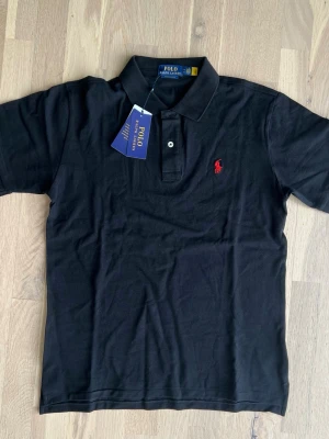 Ralph Lauren black polo shirt size M - Brand new, never worn Polo Ralph Lauren Custom Slim Fit polo shirt in Black. Comes with original tags attached and original packaging.  * Condition: Brand New with Tags (NWT) * Color: Black * Fit: Custom Slim Fit * Size: M * Material: 100% Cotton * No flaws, stains, holes, or signs of wear. * Authentic Polo Ralph Lauren. * Retail ≈ 180$ A timeless wardrobe essential, perfect for both casual and smart-casual outfits.