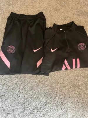 Psg tracksuit  - Snyggt svart träningsset från PSG och Nike med rosa detaljer. Setet består av långärmad tröja med halv dragkedja och matchande byxor, båda med PSG-logga och Nike swoosh i rosa. Materialet är lätt och andas, perfekt för träning eller chill.