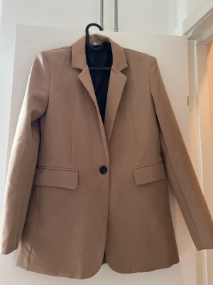 Beige/brun oversized kavaj med knapp - Stilren beige kavaj i oversized modell med en knapp framtill och två fickor med lock. Klassisk krage och svart innerfoder ger en clean look. Perfekt för att lyfta vilken outfit som helst.