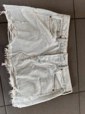 Ljus jeanskjol med fransig kant - Säljer en ljusblå jeanskjol med rå, fransig kant och slitna detaljer. Kjolen har klassisk femficksdesign och knappgylf. Perfekt för dig som gillar en avslappnad och trendig stil.