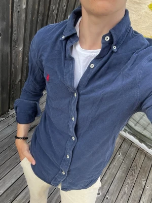 Blå skjorta från Ralph Lauren - Säljer en blå skjorta från Ralph Lauren i mjuk bomull med klassisk button-down krage och vita knappar. Skjortan har en röd broderad logga på bröstet och en avslappnad passform. Perfekt till jeans eller chinos för en clean look.
