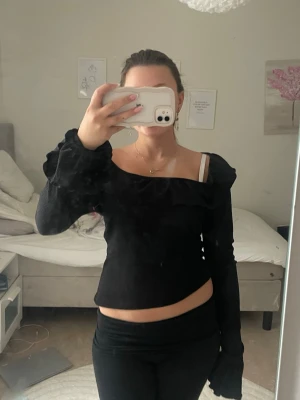 Svart långärmad topp med volang - Trendig svart långärmad topp med volangdetalj längs ringningen och ärmsluten. Toppen har en offshoulder-design och är tillverkad i ett ribbat material som ger en snygg passform. Perfekt för att lyfta din outfit med en cool touch.