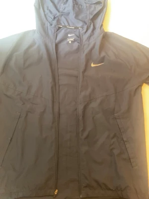 Svart vindjacka från Nike med huva - Snygg svart vindjacka från Nike med gul logga på bröstet. Jackan har dragkedja framtill, två sidofickor och en praktisk huva. Tillverkad i lätt och vindtätt material som passar perfekt för blåsiga dagar.