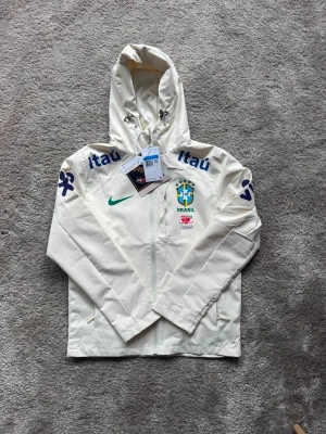 Brasilien Nike vindjacka  - Snygg vit vindjacka från Nike med Brasiliens landslagslogga på bröstet, grönt swoosh-tryck och flera sponsorloggor. Jackan har hel dragkedja, huva med dragsko och tryck på ärmarna. Perfekt för dig som gillar fotboll och sportig stil samt mode, säljs i nyskick, den var för liten för mig. Stl M men passar mer åt S hållet 