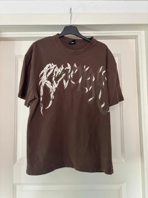 Brun t-shirt med tryck och 999-rygg - Säljer en brun t-shirt med coolt vitt tryck framtill och stora vita fåglar samt röd '999' text på ryggen. T-shirten har normal passform och är kortärmad, perfekt för dig som gillar streetwear vibes.