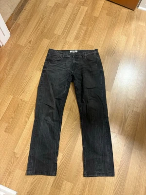 Svarta jeans från Smog Denim - Säljer ett par svarta jeans från Smog Denim i rak modell. Jeansen har klassisk femficksdesign och är tillverkade i denim med lätt stretch. Perfekta för dig som gillar en enkel och stilren look.