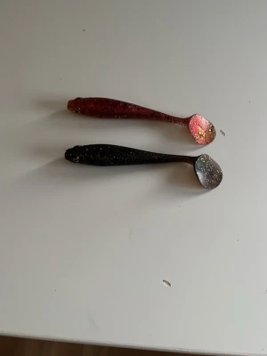 Två glittriga fiskedrag i gummi - Säljer två mjuka fiskedrag i gummi, en i svart med glitter och en i röd med glitter. Båda har fiskform och paddelstjärt, perfekta för gäddfiske eller abborre. Snygga färger som lockar fisken till hugg.