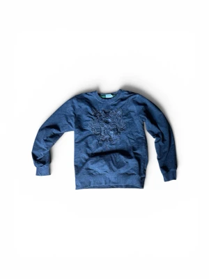Mörkgrå Kenzo sweatshirt med tiger - Mörkgrå sweatshirt från Kenzo med det ikoniska tigermotivet broderat på bröstet och texten 'KENZO PARIS'. Tröjan har rund halsringning, långa ärmar och ribbade muddar. Perfekt för en trendig och avslappnad stil. Storlek L i barn.