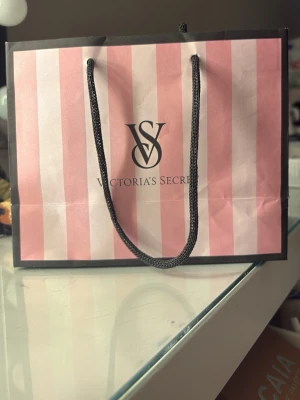 Victoria's Secret presentpåse rosa - Säljer en ikonisk presentpåse från Victoria's Secret med rosa och ljusrosa ränder samt svart logga. Påsen har svarta snören som handtag och är perfekt för att ge bort presenter eller använda som accessoar.