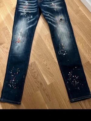 Mörkblå jeans med färgstänk - Säljer ett par mörkblå jeans med coola färgstänk och slitningar på flera ställen. Jeansen har en smal passform och klassisk femficksdesign. Perfekt för dig som gillar en unik och trendig look.