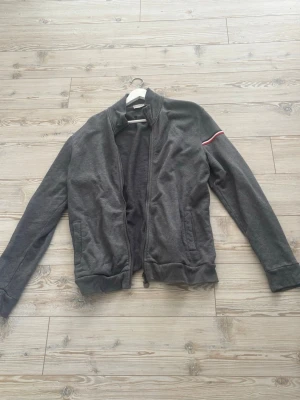 Mörkgrå zip hoodie från Moncler - Snygg mörkgrå hoodie från Moncler i storlek XL men passar L. Dragkedja framtill, två fickor och ribbade muddar. Detalj med röd och vit rand på ena ärmen. 