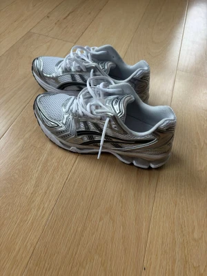 Vita och silver Asics sneakers Gel kayano 14 - Snygga vita sneakers från Asics med silvriga detaljer och svart logga på sidan. Skorna har en sportig design med mesh- och syntetmaterial för bra andningsförmåga. Klassisk snörning och vadderad insida för extra komfort.