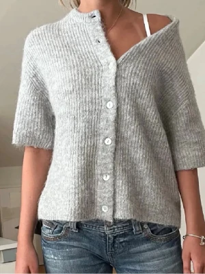Ljusgrå kofta från Gina Tricot - Mysig ljusgrå kofta från Gina Tricot i ribbstickad kvalitet. Koftan har korta ärmar, rund halsringning och knäpps med vita knappar framtill. Perfekt att slänga över ett linne eller t-shirt för en avslappnad look.