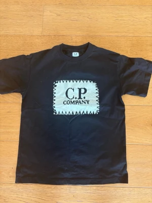 Svart C.P. Company t-shirt - Snygg svart t-shirt från C.P. Company med ett stort vitt tryck på bröstet och rund hals. T-shirten har klassisk passform och korta ärmar. Perfekt för dig som gillar streetwear och vill ha en enkel men stilren look.