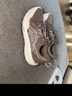Asics sneakers - Säljer ett par snygga sneakers i brunt med vita detaljer och mönstrade skosnören. Skorna har en tjock vit sula och är tillverkade i mesh och syntetmaterial. Perfekta för dig som gillar sportig stil och vill ha bekväma skor till vardagen.