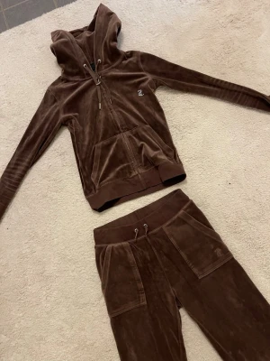 Brunt set från Juicy Couture - Säljer ett brunt set från Juicy Couture med hoodie och byxor. Hoodien har dragkedja, fickor och Juicy i strass på ryggen. Byxorna har resårmidja med snörning och fickor med knappar bak. Perfekt för chill dagar hemma.
