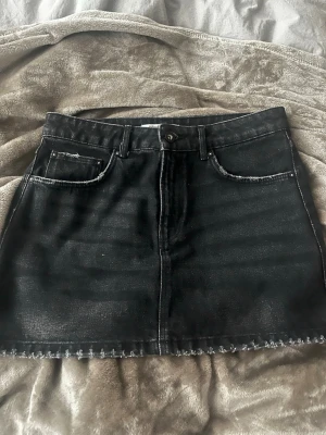 Svart jeanskjol - Snygg svart jeanskjol i kort modell, fransig kant nertill. Kjolen har klassiska fem fickor, bälteshällor och knappstängning framtill. Perfekt till en avslappnad och trendig look.