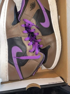Bruna och svarta sneakers med lila detaljer - Snygga sneakers i brunt och svart med lila swoosh, lila skosnören och lila detaljer på plösen. Skorna har en klassisk siluett med högt skaft och vit sula. Perfekta för dig som vill sticka ut med färg och stil.