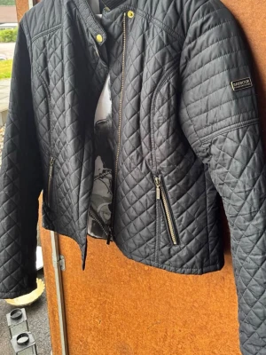 Svart quiltad jacka Barbour - Snygg svart quiltad jacka från Barbour med gulddetaljer och ståkrage. Jackan har två dragkedjefickor framtill och ett coolt innerfoder med motorcykeltryck. Perfekt för dig som gillar klassisk men edgy stil.