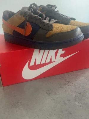 Nike Dunk Low sneakers brun/orange - Nike Dunk Low sneakers med brun mocka, mörkblå och grå detaljer samt orange swoosh. Skorna har klassisk låg siluett, perforerad tå och grå snörning. Snygg färgkombo som sticker ut och passar perfekt till streetwear.