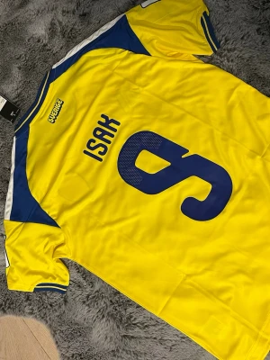 Alexander Isak sverige tröja - Gul och blå Sverige fotbollströja från Adidas med nummer 9 och ISAK på ryggen. Tröjan har blå detaljer på axlar och ärmar, samt officiella landslagsmärket på bröstet. Tillverkad i lätt och ventilerande material, perfekt för match eller träning.