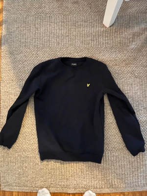 Mörkblå sweatshirt från Lyle & Scott - Mörkblå sweatshirt från Lyle & Scott med klassisk rund halsringning och den ikoniska gula örnloggan broderad på bröstet. Tröjan har långa ärmar och ribbade muddar vid ärmslut och nederkant. Perfekt för en avslappnad och stilren look.
