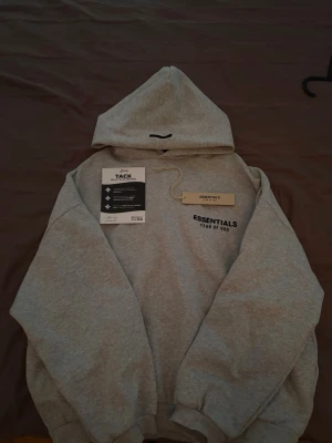 Grå hoodie från Essentials Fear of God - Snygg grå hoodie från Essentials Fear of God med svart logga på bröstet. Tillverkad i mjuk bomullsblandning (80% bomull, 20% polyester) och har en klassisk huva samt ribbade muddar. Perfekt för en avslappnad streetwear-look.