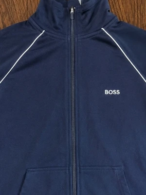 Mörkblå zip-hoodie från BOSS - Mörkblå hoodie med dragkedja från BOSS. Den har vita kontrastlinjer längs axlarna och en liten BOSS-logga på bröstet. Praktiska fickor framtill och klassisk huva. Perfekt för en avslappnad och stilren look.
