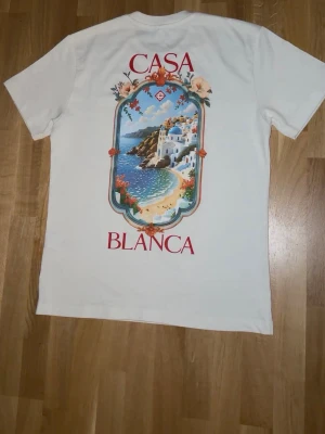Vit Casa Blanca t-shirt - Vit t-shirt från Casa Blanca med färgglatt tryck av ett medelhavsmotiv på ryggen och mindre tryck framtill. T-shirten har rund hals och korta ärmar. Perfekt för dig som gillar stilrena plagg med unik design.