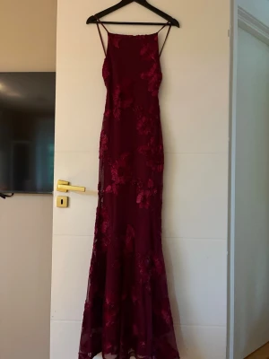 Vinröd långklänning med spetsdetaljer - Elegant vinröd långklänning med tunna axelband och blommiga spetsdetaljer. Klänningen har figurnära passform och är perfekt för speciella tillfällen. Tunn, lätt genomskinlig överdel med vackert fall.