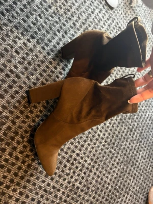 Bruna stretchiga stövletter med klack - Säljer ett par bruna stövletter med hög, bred klack och spetsig tå. Skorna är i stretchigt tyg som sitter tight runt ankeln och har en modern siluett. Perfekta för dig som vill ha en trendig look med bekväm passform.