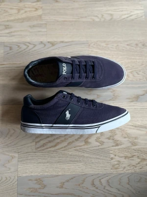 Polo Ralph Lauren sneakers - Navy Canvas, 44 - Bra skick. Se små defekter nedan.  Canvas-sneakers från Polo Ralph Lauren, storlek 44, marinblå Stilrena skor. Blåa med vit sula, logga på yttersidan samt texten “POLO” vid snörning.  Ovandel: 71% canvas, 29% PU (syntetiskt läder) Foder: 79% textil, 21% läder  Yttersula: 100% gummi  OBS: Delvis bortfall av texten “POLO” i hälen på ena skon. Innersulor saknas.