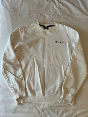 Vit sweatshirt från Champion - Klassisk vit sweatshirt från Champion med rund halsringning och diskret broderad logga på bröstet. Tröjan har långa ärmar och ribbade muddar vid ärmslut och nederkant. Perfekt för en clean och avslappnad stil.