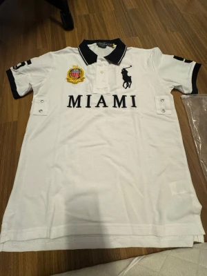 Polo ralph lauren - Miami - Vit pikétröja från Polo Ralph Lauren med svart krage och detaljer. Stor MIAMI-text framtill, broderad logga och emblem på bröstet. Klassisk polokrage med knappar och korta ärmar med svarta muddar. Perfekt för en sportig och stilren look. Slim fit