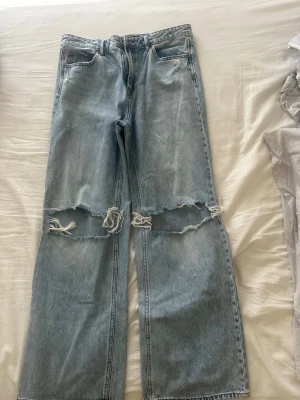 Ljusblå ripped jeans Stradivarius - Säljer ett par ljusblå jeans från Stradivarius i storlek 42. Modellen har raka ben och stora slitningar över knäna för en trendig, avslappnad look. Klassisk femficksdesign och tillverkade i bomullsdenim. Perfekta för dig som gillar streetstyle.