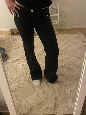 Svarta bootcut jeans med silverknappar - Säljer ett par svarta bootcut jeans med snygga silverfärgade knappar framtill. Jeansen har låg midja och klassisk femficksdesign. Materialet ser ut att vara stretchigt denim som sitter bekvämt och formar sig efter kroppen.