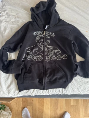 Svart hoodie med tryck och dragkedja - Svart hoodie med huva och dragkedja framtill. På framsidan finns ett stort vitt tryck med text och en grafisk illustration av en person. Hoodien har fickor på magen och ribbade muddar vid ärmar och nederkant. Perfekt för en avslappnad streetwear-look.
