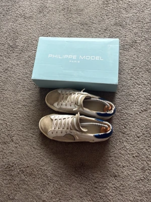 Philippe Model vita sneakers - Snygga vita sneakers från Philippe Model med beige mockadetaljer på tån och blå detalj på hälen. Klassisk låg modell med vita snören och gummisula. Perfekta för dig som gillar stilrena och trendiga skor. Självklart äkta, Box tillkommer även.
