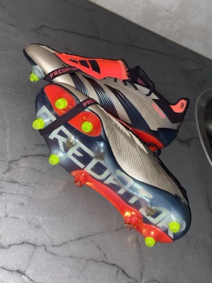 Adidas Predator fotbollsskor silver/röd - Snygga Adidas Predator fotbollsskor i silver med röda och svarta detaljer. Skorna har en modern design med texten 'Predator' på sulan och neongula dobbar för grymt grepp på planen. Ovandelen är i syntetmaterial och insidan är röd för extra stilpoäng.