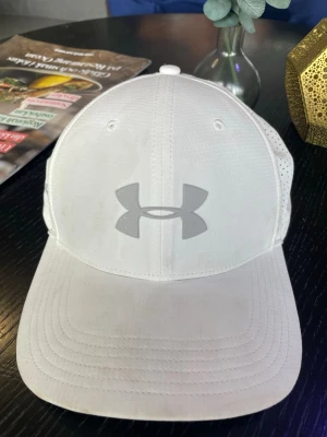 Vit keps från Under Armour - Snygg vit keps från Under Armour med ventilerande meshpaneler på sidorna och böjd skärm. Stor logga framtill och justerbar passform bak. Perfekt för en sportig och fräsch look.