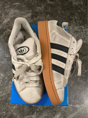 Adidas campus  - Snygga beige sneakers från Adidas med klassiska svarta ränder på sidan och tjock gummisula i brun färg. Ovandelen är i mockaliknande material och skorna har låga skaft. Perfekta för en streetwear-look.