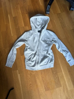 Grå hoodie från Peak Performance - Snygg ljusgrå hoodie från Peak Performance med dragkedja och broderad logga på bröstet. Mjuk bomullsblandning, två fickor framtill och klassisk huva. Perfekt för en avslappnad och sportig stil. OBS! Storlek 150 i barn!