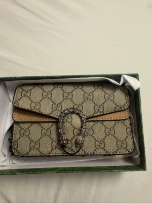 GG supreme handväska - Säljer en handväska i beige med bruna detaljer och silverfärgat dekorativt spänne framtill. Perfekt för dig som gillar lyxiga accessoarer och tidlös design.