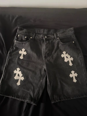 Svarta Chrome hearts Denim shorts med korsdetaljer - Unika svarta shorts i denim med stora vita korsapplikationer både fram och bak. Klassisk femficksmodell med normal passform och bälteshällor. Perfekta för dig som vill sticka ut med en edgy streetwear-look.