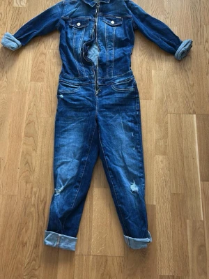 Blå denim jumpsuit från Guess - Snygg blå denim jumpsuit från Guess med dragkedja framtill, bröstfickor med knappar och upprullade ärm- och benslut. Tuffa slitningar på benen ger en cool look. Perfekt för dig som gillar streetstyle och denim.