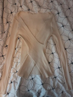 Beige ribbad omlottopp - Säljer en beige ribbad tröja med omlottdesign och långa ärmar. Tröjan har en figurnära passform och är tillverkad i ett stretchigt material som ger en snygg siluett. Perfekt för dig som gillar stilrena och trendiga plagg.
