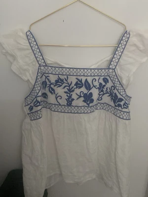 Vit blus med blå broderi och knyt - Söt vit blus med breda axelband och blått broderat blommönster framtill. Toppen har knyt i ryggen och volangdetaljer vid ärmarna. Perfekt för en somrig och avslappnad stil.