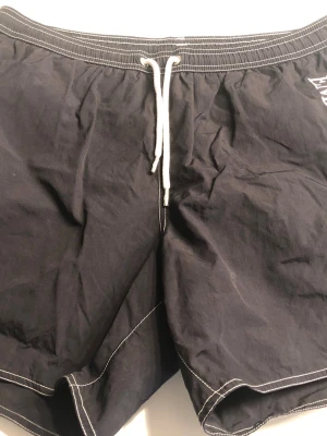 Marinblåa badbyxor Emporio Armani - Snygga marinblåa badbyxor från Emporio Armani med vit dragsko i midjan och vit kontrastsöm. Stort Emporio Armani-tryck längs ena benet. Klassisk passform och snabbtorkande material, perfekta för stranden eller poolen.